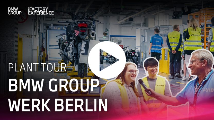 Header Plant Tour BMW Group Werk Berlin, Guide hält Besucherin ein Mikrofon hin, im Hintergrund sind weitere Besuchende und Motorräder zu sehen